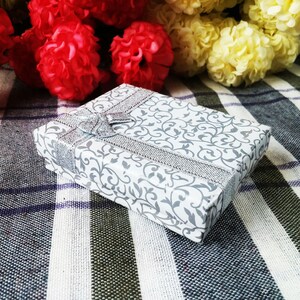 May include: A white gift box with a silver ribbon and a floral pattern. The box is sitting on a checkered fabric.