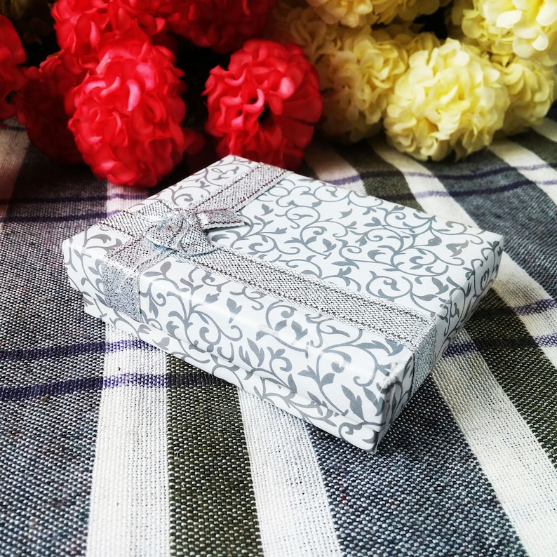May include: A white gift box with a silver ribbon and a floral pattern. The box is sitting on a checkered fabric.