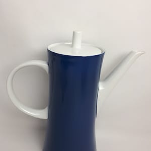 Puede incluir: Una cafetera de cerámica blanca y azul con una tapa blanca y un pico. La cafetera tiene una forma alta y cilíndrica y una asa blanca.