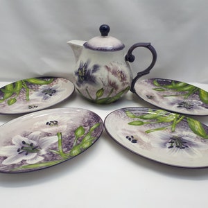 Puede incluir: Un juego de cuatro platos ovalados blancos con un diseño floral morado y verde y una tetera a juego con tapa morada.