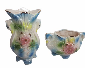 May include: A pair of ceramic vases featuring a swan design. The vases are decorated with blue, green, yellow and pink floral patterns. The larger vase has a wide mouth and a narrow base. The smaller vase is more rounded and has a wider base.
