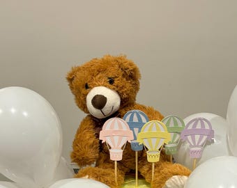 May include: A brown teddy bear sits surrounded by white balloons and colorful hot air balloon decorations. The balloons are on sticks and have a pastel color scheme.