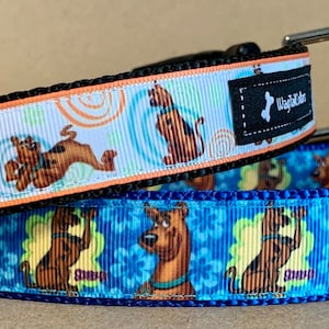 Puede incluir: Dos collares para perros con temática de Scooby-Doo. El collar superior es blanco con remolinos naranjas y Scooby-Doo corriendo. El collar inferior es azul con flores amarillas y Scooby-Doo de pie. Ambos collares tienen una correa negra y una hebilla plateada.
