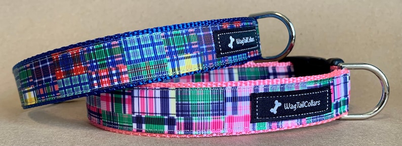 May include: Two dog collars with a plaid pattern. One collar is blue with red, green, and yellow accents. The other collar is pink with green, purple, and white accents. Both collars have a metal D-ring and a black label that says "WagTailCollars".