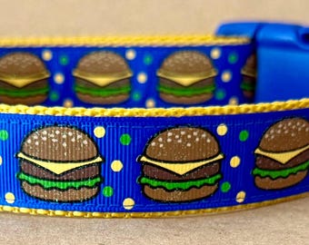 May include: A blue pet collar featuring a repeating pattern of cheeseburgers and small green and yellow polka dots. The collar has a yellow fabric trim, a blue plastic side-release buckle, a silver metal D-ring, and a small black rectangular label.