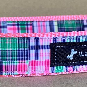 May include: A dog collar with a pink trim and a colorful plaid pattern in shades of pink, green, and navy blue. The collar has a black buckle and a metal D-ring. The label reads "WagTailCollars".