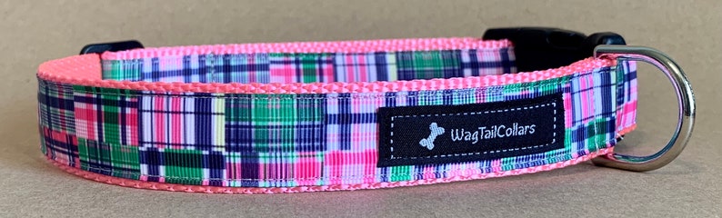 May include: A dog collar with a pink trim and a colorful plaid pattern in shades of pink, green, and navy blue. The collar has a black buckle and a metal D-ring. The label reads "WagTailCollars".