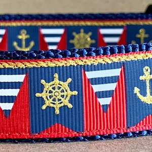 Pode incluir: Coleira azul marinho para cães com tema náutico. A coleira apresenta bandeirolas vermelhas e brancas, âncoras e rodas de navio douradas, e o texto "WagTail Collars" em uma etiqueta preta. Possui um anel em D de metal e uma fivela de plástico preta.
