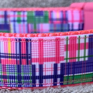 May include: A colorful dog collar with a patchwork plaid pattern in pink, green, blue, and white. The collar has a pink plastic buckle and a metal D-ring. The inner lining is a solid pink, and a black tag with white text is visible.