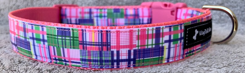 May include: A colorful dog collar with a patchwork plaid pattern in pink, green, blue, and white. The collar has a pink plastic buckle and a metal D-ring. The inner lining is a solid pink, and a black tag with white text is visible.