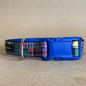 May include: A blue dog collar with a plaid pattern in red, green, and yellow. The collar has a blue plastic buckle and a silver metal ring. The brand name is printed on a black label.
