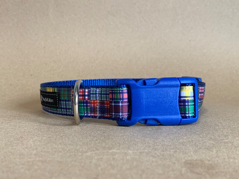 May include: A blue dog collar with a plaid pattern in red, green, and yellow. The collar has a blue plastic buckle and a silver metal ring. The brand name is printed on a black label.