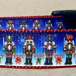Könnte beinhalten: Ein Haustierhalsband mit einem sich wiederholenden Muster aus Nussknacker-Figuren auf blauem Grund mit weißen Schneeflocken. Das Halsband verfügt über eine schwarze Kunststoff-Steckschnalle, einen silbernen Metall-D-Ring und einen dunkelroten Stoffrand.