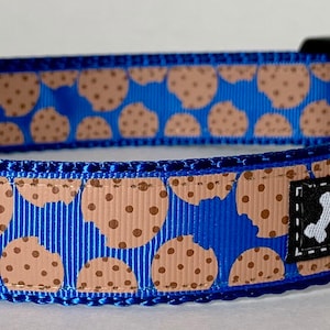 Puede incluir: Collar azul para perros con un estampado de galletas color beige con chispas de chocolate marrón. El collar tiene una hebilla de plástico negro y una anilla en D de metal plateado. La marca "Wag Tail Collars" está en una etiqueta negra con un hueso blanco.