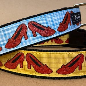 Pode incluir: Duas coleiras para cães com um design de caminho de tijolos amarelos e sapatos vermelhos de rubi. A coleira superior é azul e branca xadrez. A coleira inferior é amarela com um padrão de caminho de tijolos.