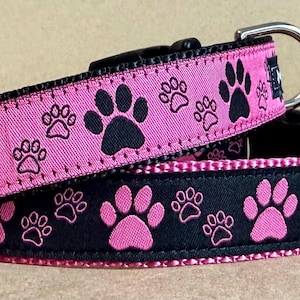 May include: Two dog collars, one pink and one black, each decorated with paw prints. The pink collar has black paw prints, while the black collar has pink paw prints. Both collars have a silver metal D-ring and a black plastic buckle.