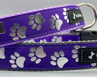 May include: Two purple dog collars with silver paw prints. Each collar has a black plastic buckle, a metal D-ring, and a label that reads "Wag Tail Collars". The collars are stacked on top of each other.