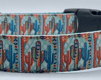 May include: A dog collar with a Southwestern-inspired design featuring cacti in shades of blue, orange, and green. The collar has a black plastic buckle, a silver metal D-ring, and a label that reads "Wag Tail Collars". The interior is teal.