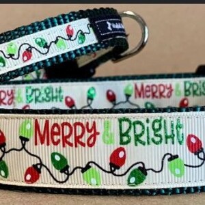May include: Two dog collars with a white background and green and red Christmas lights. The collars have the text "Merry & Bright" on them. The larger collar has a black buckle and a silver D-ring. The smaller collar has a black buckle and a silver loop.