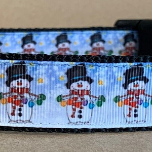 May include: A black dog collar with a repeating pattern of snowmen wearing top hats and holding colorful lights against a light blue background. The collar has a black buckle and a metal D-ring.