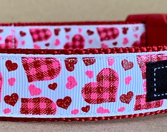 May include: Red dog collar with a white background and a pattern of pink and red plaid hearts. The collar has a silver D-ring and a red plastic buckle. The collar has a black label that says "WagTailCollars" with a bone icon.