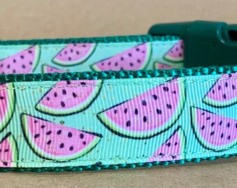 May include: A pet collar featuring a repeating pattern of pink watermelon slices on a light mint green background. The collar includes a dark green plastic side-release buckle, a metal D-ring, and a small black fabric tag labeled WagTailCollars.