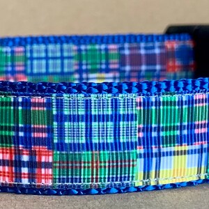 May include: A blue dog collar with a patchwork plaid pattern in red, green, yellow, and white. The collar has a black buckle, a silver metal D-ring, and a label that says "WagTailCollar".