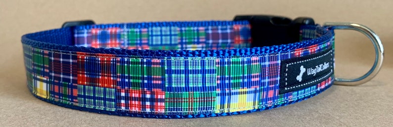 May include: A blue dog collar with a patchwork plaid pattern in red, green, yellow, and white. The collar has a black buckle, a silver metal D-ring, and a label that says "WagTailCollar".
