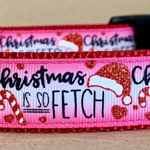 Puede incluir: Un collar rosa para perros con el texto "Christmas is so FETCH" en escritura negra. El collar presenta gorros de Papá Noel, bastones de caramelo y corazones. El collar tiene una hebilla negra y una anilla en D de metal plateado.