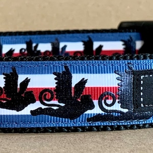 May include: A blue, white, and red dog collar with a black bird silhouette pattern. The collar has a black buckle and a silver metal D-ring. The brand name "WagTailColors" is displayed on a black patch.