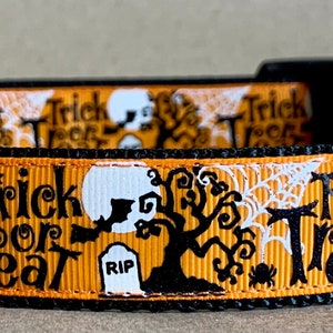 May include: Halloween-themed dog collar with a black base and orange ribbon featuring white skulls, spiderwebs, trees, and the words "Trick or Treat." The collar has a black plastic buckle and a silver metal D-ring.