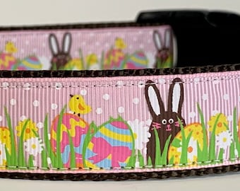 May include: Pink dog collar with a brown border and an Easter-themed pattern featuring bunnies, eggs, and chicks. The collar has a silver buckle and a black tag that says "Wag'n Tails".