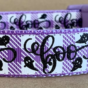Op de afbeelding: Een paars en wit geruite halsband voor huisdieren met een patroon van zwarte spookjes en het woord boo in cursief schrift. De halsband is voorzien van een lichtpaarse plastic gesp, een metalen D-ring en een zwart label met de tekst WagTailCollars.