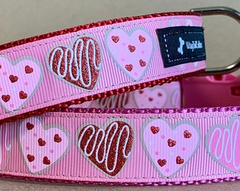May include: Two pink dog collars with a red and white heart pattern. The collars have a silver D-ring and a black tag that says "Puplife".