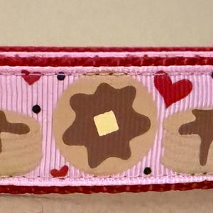 May include: Pink dog collar with a red border and a silver D-ring. The collar features a repeating pattern of brown pancakes with a yellow pat of butter and red hearts.