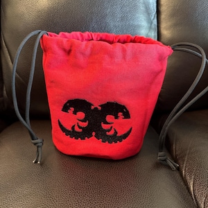 May include: A bright red drawstring pouch made of canvas fabric. The bag features a black embroidered graphic design on the front, consisting of two mirrored, stylized circular shapes. It has two black cord drawstrings on the sides.