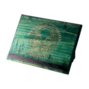 Könnte beinhalten: Grüner hölzerner Buchständer mit den Worten "Miskatonic University, Arkham, MA" und einem kreisförmigen Emblem in Gold geätzt. Der Ständer ist zum Präsentieren abgewinkelt und hat eine dunkelbraune Kante.