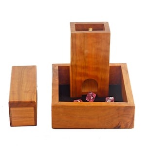 May include: Wooden dice tower with a removable tray and a separate dice box. The tower has a slot for dice to fall through into the tray. The tray is lined with black felt and has dice in it. The dice are pink and have white numbers.