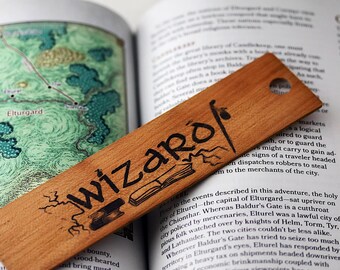 Puede incluir: Un marcapáginas de madera con la palabra "wizaro" grabada en letras negras. El marcapáginas está sobre una página de un libro abierto con un mapa y texto sobre la ciudad de Elturgard.