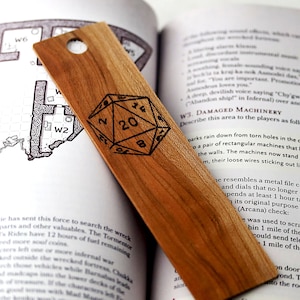 Könnte beinhalten: Ein Holz-Lesezeichen mit einem schwarz-weißen Bild eines 20-seitigen Würfels. Das Lesezeichen ist mit den Worten "20" und "D20" graviert.