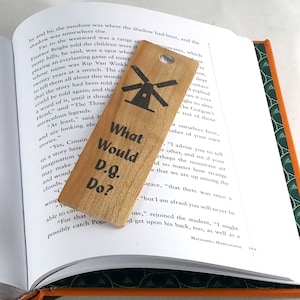 May include: Wooden bookmark with a windmill illustration and the text "What Would D.Q. Do?" The bookmark is light brown and has a hole at the top. It is placed on an open book.
