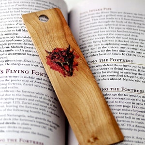 May include: A wooden bookmark featuring a red and black illustration of a dragon's head. The bookmark is made from natural wood and has a distressed finish. The text "Z'neth's Flying Fortress" is printed on the bookmark.