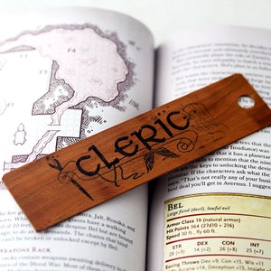 Könnte beinhalten: Ein Holz-Lesezeichen mit dem Wort "cleric" in schwarzer Tinte. Das Lesezeichen liegt auf einem geöffneten Buch mit Text über einen "Großen Dämon (Teufel), gesetzloses Böses" namens Bel. Der Text enthält die Werte des Charakters, wie Rüstungsklasse, Trefferpunkte und Geschwindigkeit.