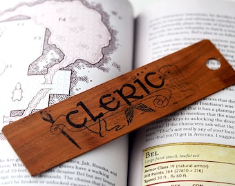 Puede incluir: Un marcapáginas de madera con la palabra "cleric" escrita en tinta negra. El marcapáginas está sobre un libro abierto con texto sobre un "Gran demonio (diablo), mal legal" llamado Bel. El texto incluye las estadísticas del personaje, como la clase de armadura, los puntos de golpe y la velocidad.