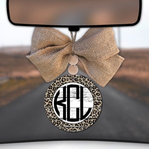 May include: A car air freshener with a burlap bow and a round wooden disc with a white and black monogram "KCL" on a woodgrain background with a leopard print border.