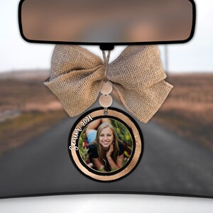 May include: A black and gold round car air freshener with a photo of a young woman and the text "Senior 2021". The air freshener is hanging from a rearview mirror with a brown burlap bow.