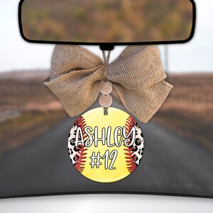 May include: A yellow softball car air freshener with a brown burlap bow. The softball has red stitching and a black and brown leopard print design. The text "Ashley #12" is printed on the softball.