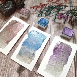 May include: Three watercolor paint pans with metallic finishes. The pans are on a wooden surface with a sprig of dried flowers. The colors are a rose gold, a blue with silver shimmer, and a purple with silver shimmer.