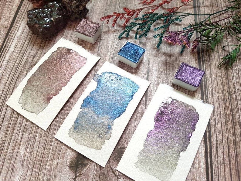 May include: Three watercolor paint pans with metallic finishes. The pans are on a wooden surface with a sprig of dried flowers. The colors are a rose gold, a blue with silver shimmer, and a purple with silver shimmer.