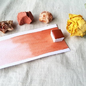 Könnte beinhalten: Eine Aquarell-Farbpalette mit einem braunen Farbmuster auf einem weißen Papierkarton. Die Farbpalette befindet sich auf einer weißen Leinenoberfläche mit einer gelben Rose, einem kleinen Stück rotem Gestein und einem kleinen Stück weißem Gestein.